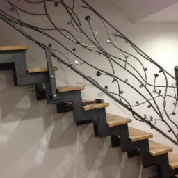 Metalowe schody z drewnianymi stopniami i ozdobną, kutą balustradą w kształcie gałęzi z liśćmi i kwiatami, widok z dołu.