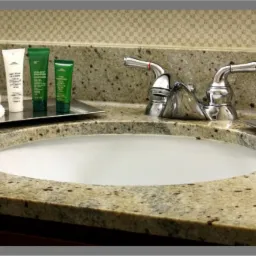 Umywalka w łazience z blatem z jasnego, nakrapianego granitu, chromowana bateria, białe mydło na metalowej podstawce i zestaw kosmetyków hotelowych na tacy.