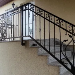 Zewnętrzne schody z szarego granitu z czarną, kutą balustradą o dekoracyjnych elementach roślinnych i geometrycznych, oświetlone lampą ścienną.