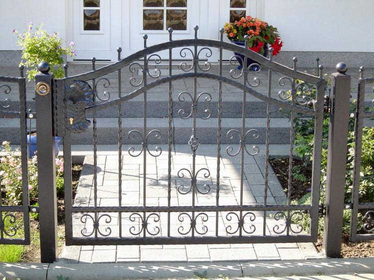 Kuta, metalowa brama wejściowa z ozdobnymi elementami w kształcie liści i kwiatów, prowadząca do domu z białymi drzwiami i schodami z kostki brukowej.