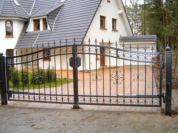 Czarna, metalowa brama wjazdowa z ozdobnymi elementami na tle białego domu z szarą dachówką i kostki brukowej.