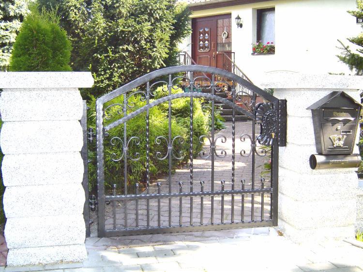 Metalowa brama wjazdowa z ozdobnymi elementami, osadzona między dwoma kamiennymi słupkami, z widoczną skrzynką pocztową i zadbaną zielenią w tle oraz fragmentem domu.