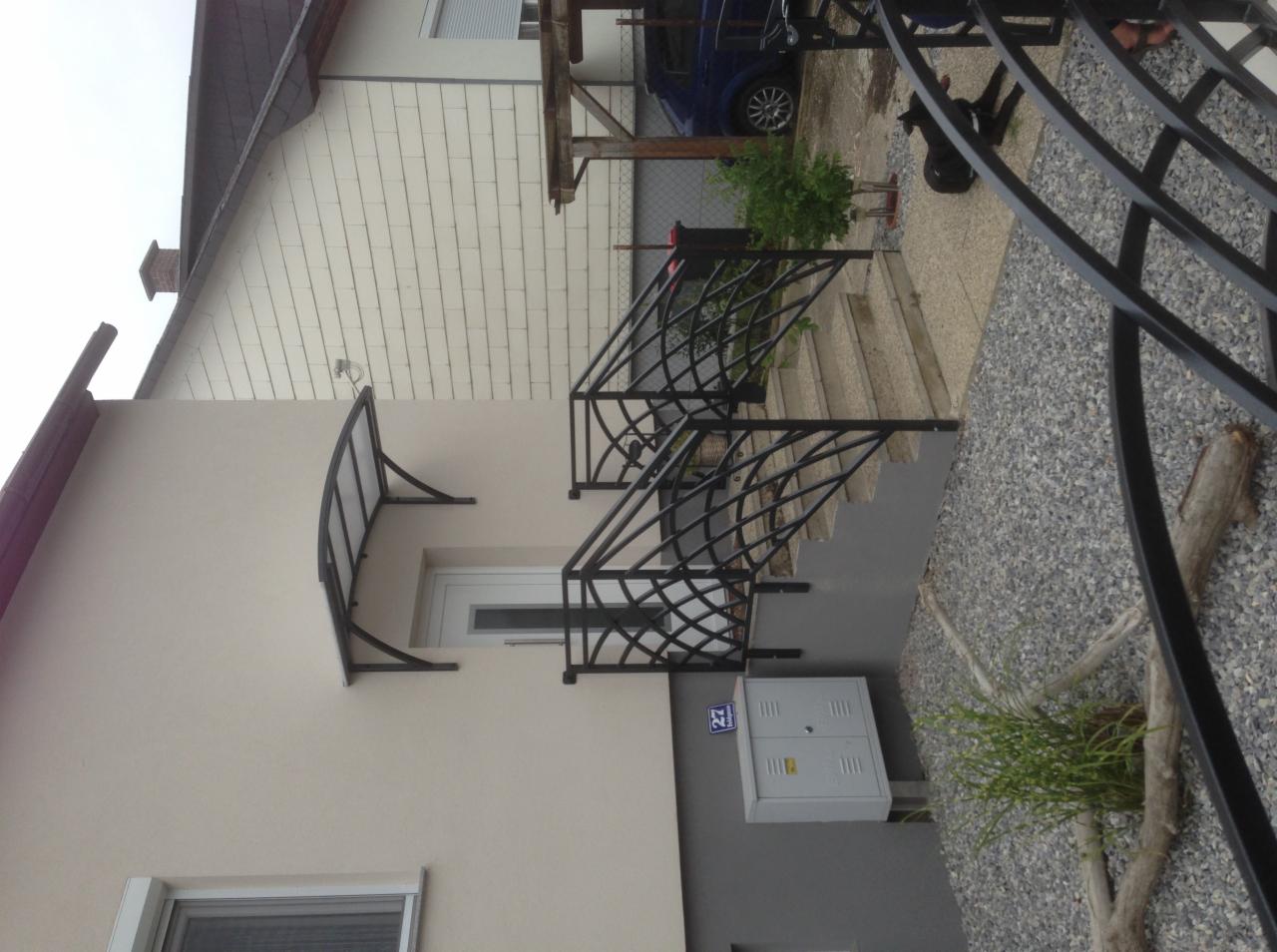 Czarna, metalowa konstrukcja schodów zewnętrznych z ozdobnym wzorem kratki, zadaszenie nad wejściem do domu, widok z boku.