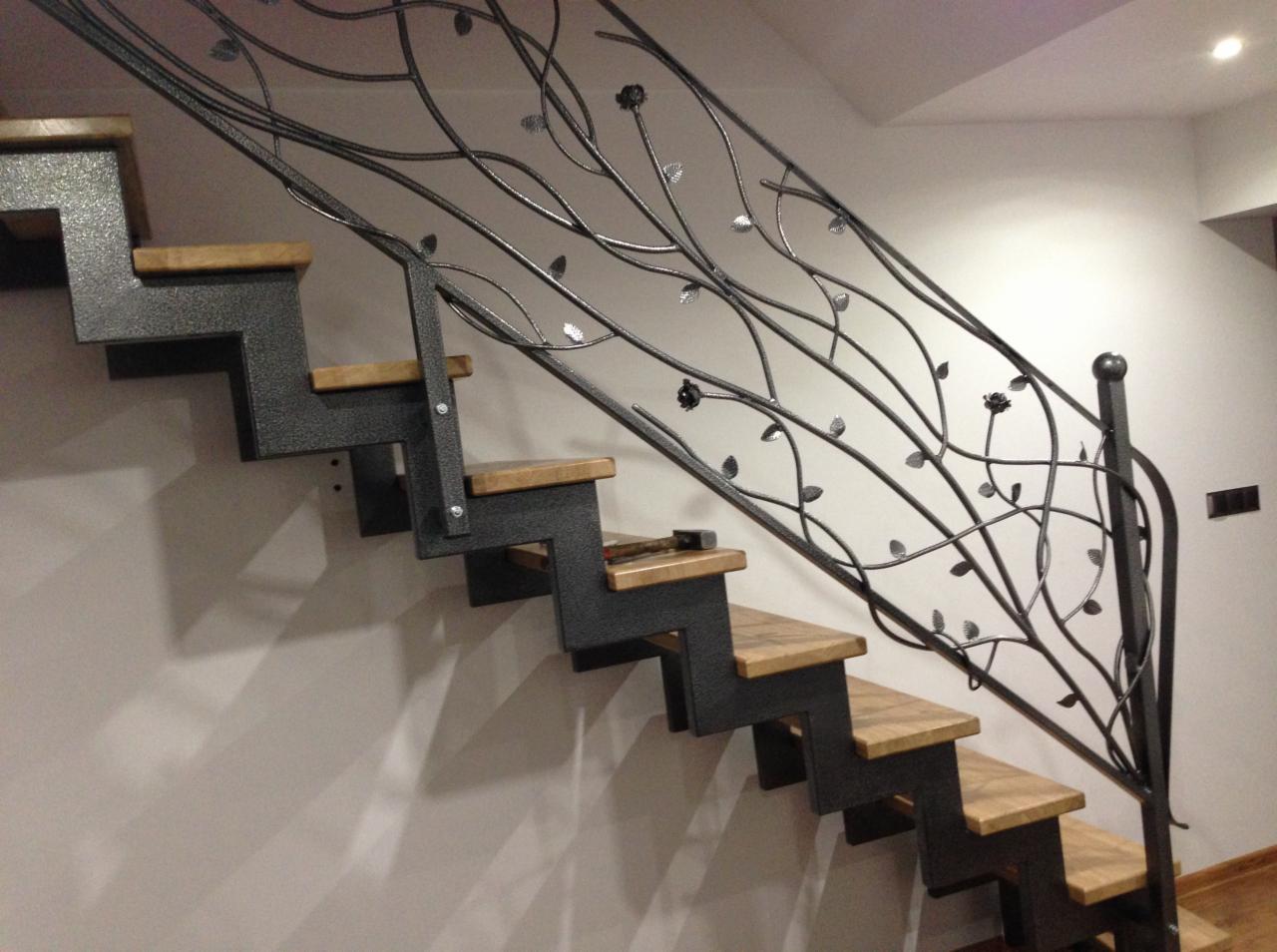 Metalowe schody z drewnianymi stopniami i ozdobną, kutą balustradą w kształcie gałęzi z liśćmi i kwiatami, widok z dołu.