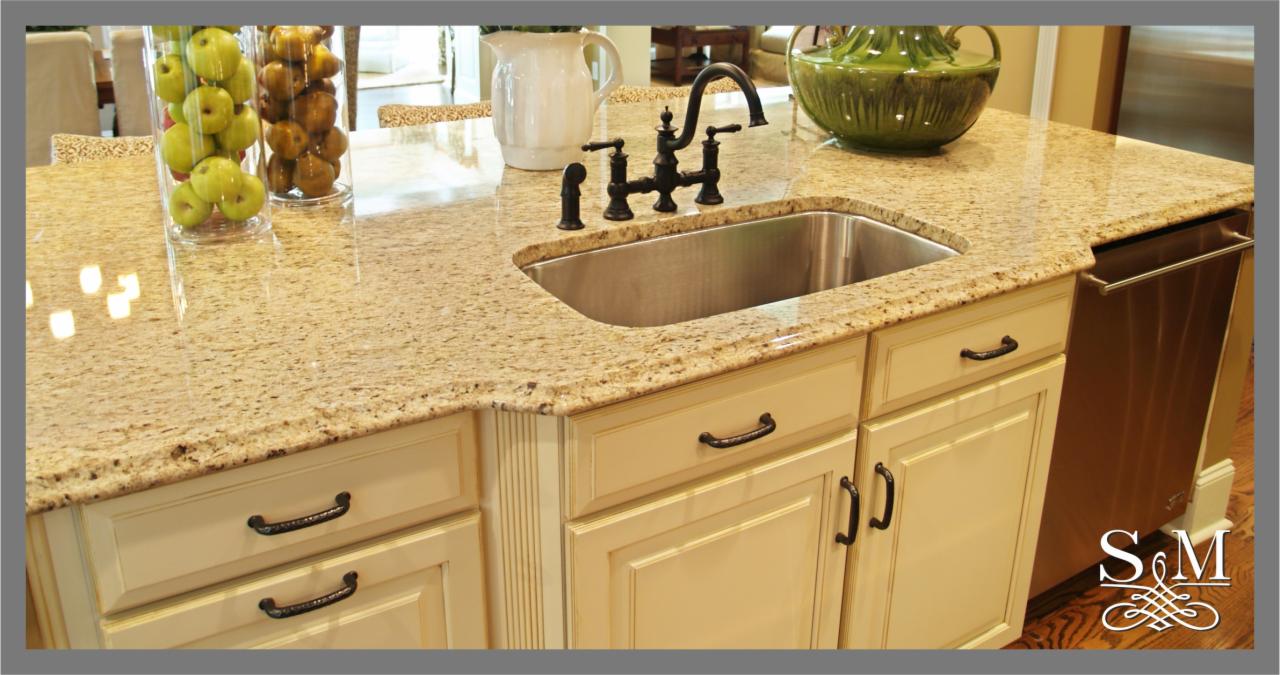 Beżowy blat kamienny z brązowymi i czarnymi plamkami na wyspie kuchennej, z czarną baterią, stalowym zlewem, szklanymi wazonami z owocami i beżowymi szafkami z czarnymi uchwytami.