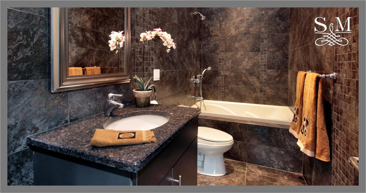 Elegancka łazienka z ciemnymi, kamiennymi płytkami, umywalką z blatem z ciemnego granitu, lustrem w srebrnej ramie i białymi akcentami w postaci wanny i toalety.