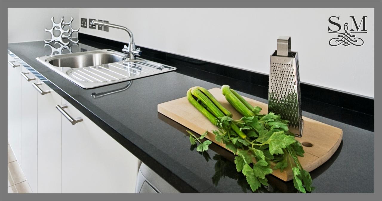 Minimalistyczna kuchnia z czarnym blatem, stalowym zlewem i świeżymi warzywami na drewnianej desce, uzupełniona o srebrną tarkę i dekoracyjny element.