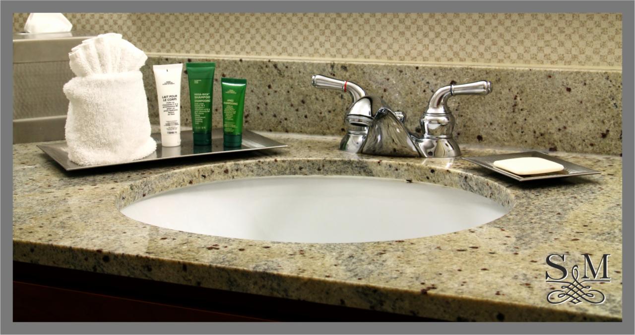 Umywalka w łazience z blatem z jasnego, nakrapianego granitu, chromowana bateria, białe mydło na metalowej podstawce i zestaw kosmetyków hotelowych na tacy.