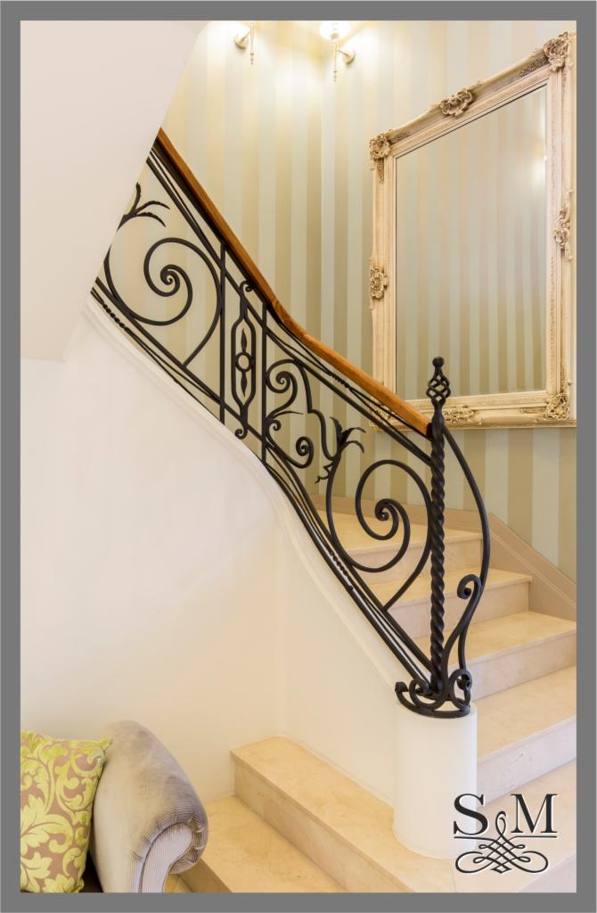 Kute, czarne balustrady z drewnianym pochwytem na marmurowych schodach w eleganckim wnętrzu z lustrem w ozdobnej ramie i tapetą w pionowe pasy.