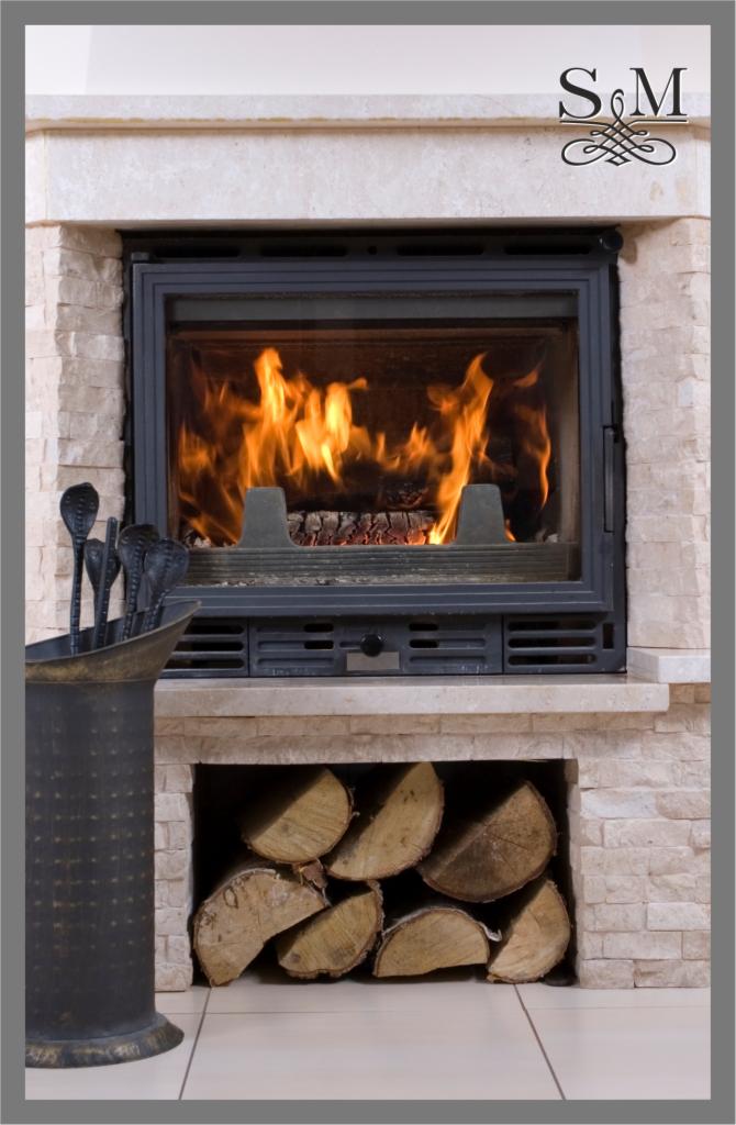 Otwarty kominek z palącym się drewnem, obudowany kamieniem w odcieniach beżu, z zestawem narzędzi kominkowych w metalowym wiadrze i zapasem drewna pod paleniskiem.