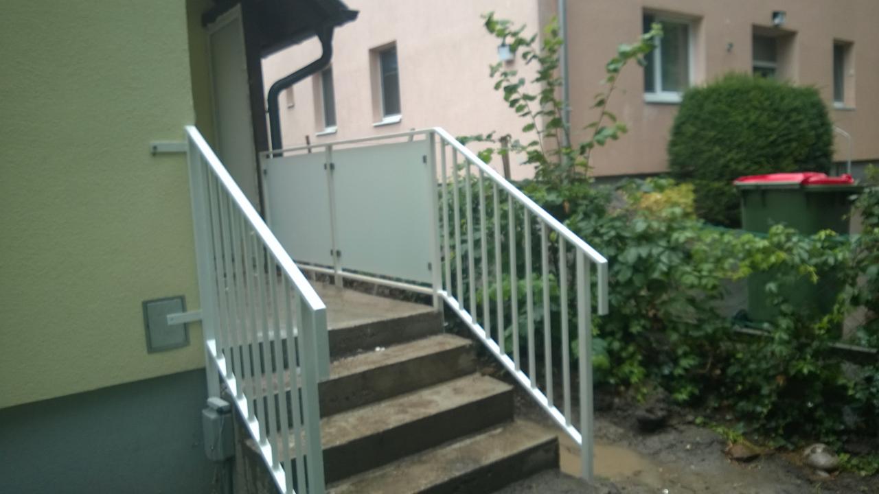 Zewnętrzne schody betonowe z białą, metalową balustradą i szklanym panelem, prowadzące do wejścia budynku mieszkalnego.