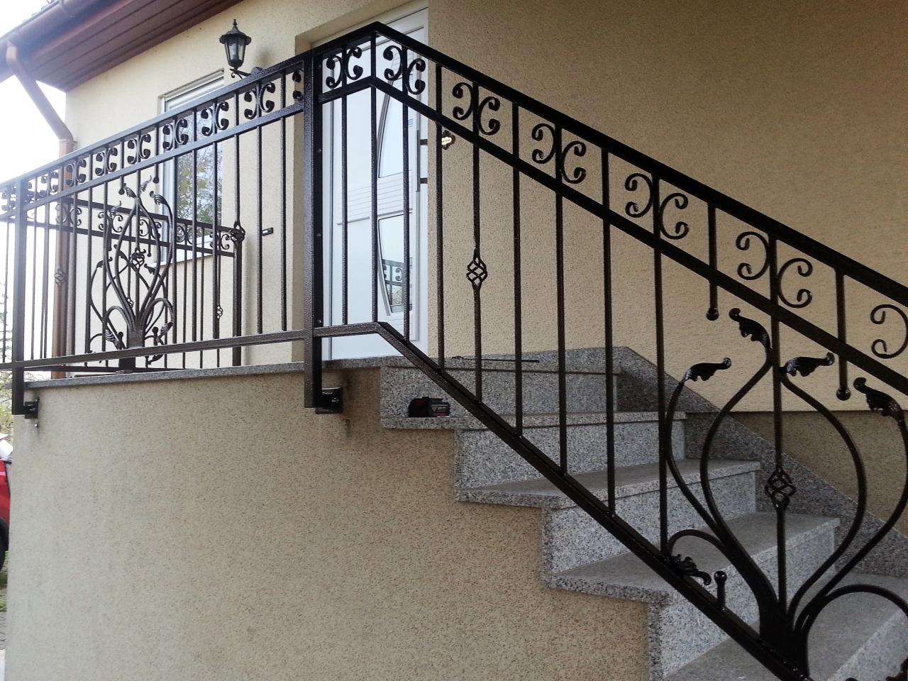 Zewnętrzne schody z szarego granitu z czarną, kutą balustradą o dekoracyjnych elementach roślinnych i geometrycznych, oświetlone lampą ścienną.