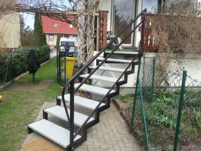 Zewnętrzne schody z czarną metalową konstrukcją i szarymi, granitowymi stopniami, prowadzące do wejścia do domu, z widocznym ogrodem i zielonym ogrodzeniem siatkowym.