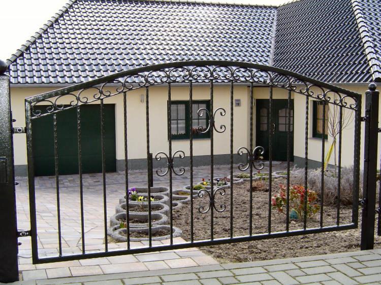 Czarna, metalowa brama wjazdowa z ozdobnymi elementami na tle domu z ciemną dachówką i zielonymi drzwiami garażowymi.