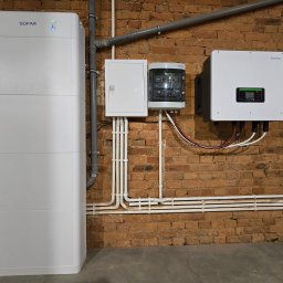 WOJ-TECH Wojciech Sidor - Instalacja pompy ciepła SOFAR na ceglanej ścianie. Widoczne białe moduły, skrzynka elektryczna i okablowanie. Elementy instalacji w pomieszczeniu z oknem.