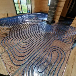 Niebieskie rury systemu ogrzewania podłogowego ułożone na folii z siatką w drewnianym domu w trakcie instalacji, widoczny fragment komina.