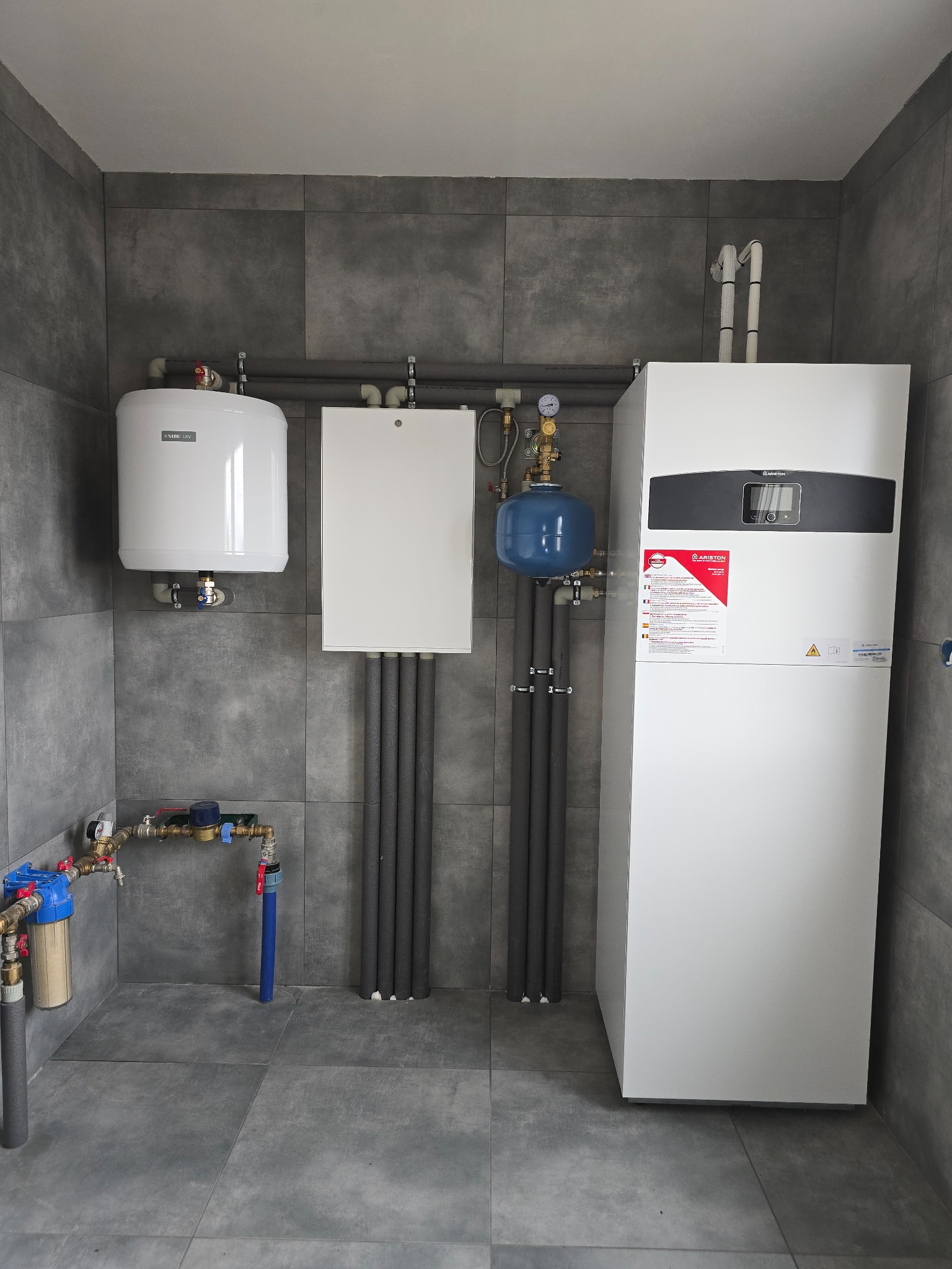 Biała instalacja grzewcza Ariston z szarymi rurami w kotłowni wyłożonej płytkami w odcieniach szarości. Widoczny bojler i filtr wody.