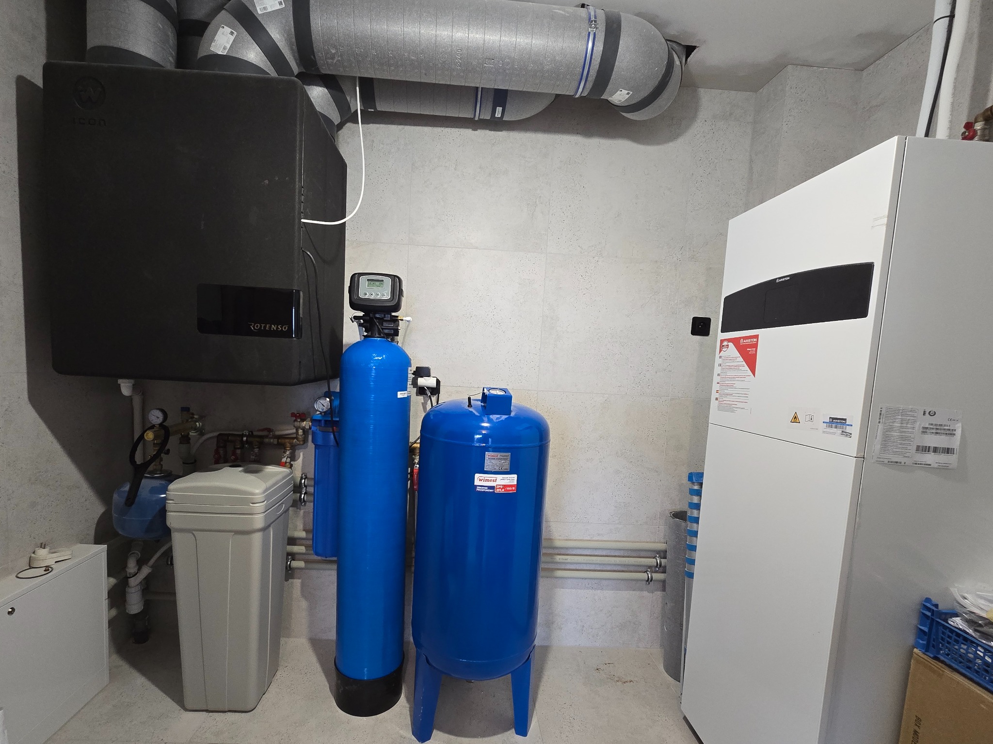 Kotłownia z czarnym piecem, niebieskimi filtrami wody i białym zasobnikiem ciepła. Widoczne rury wentylacyjne pod sufitem i szara płytka na ścianach.