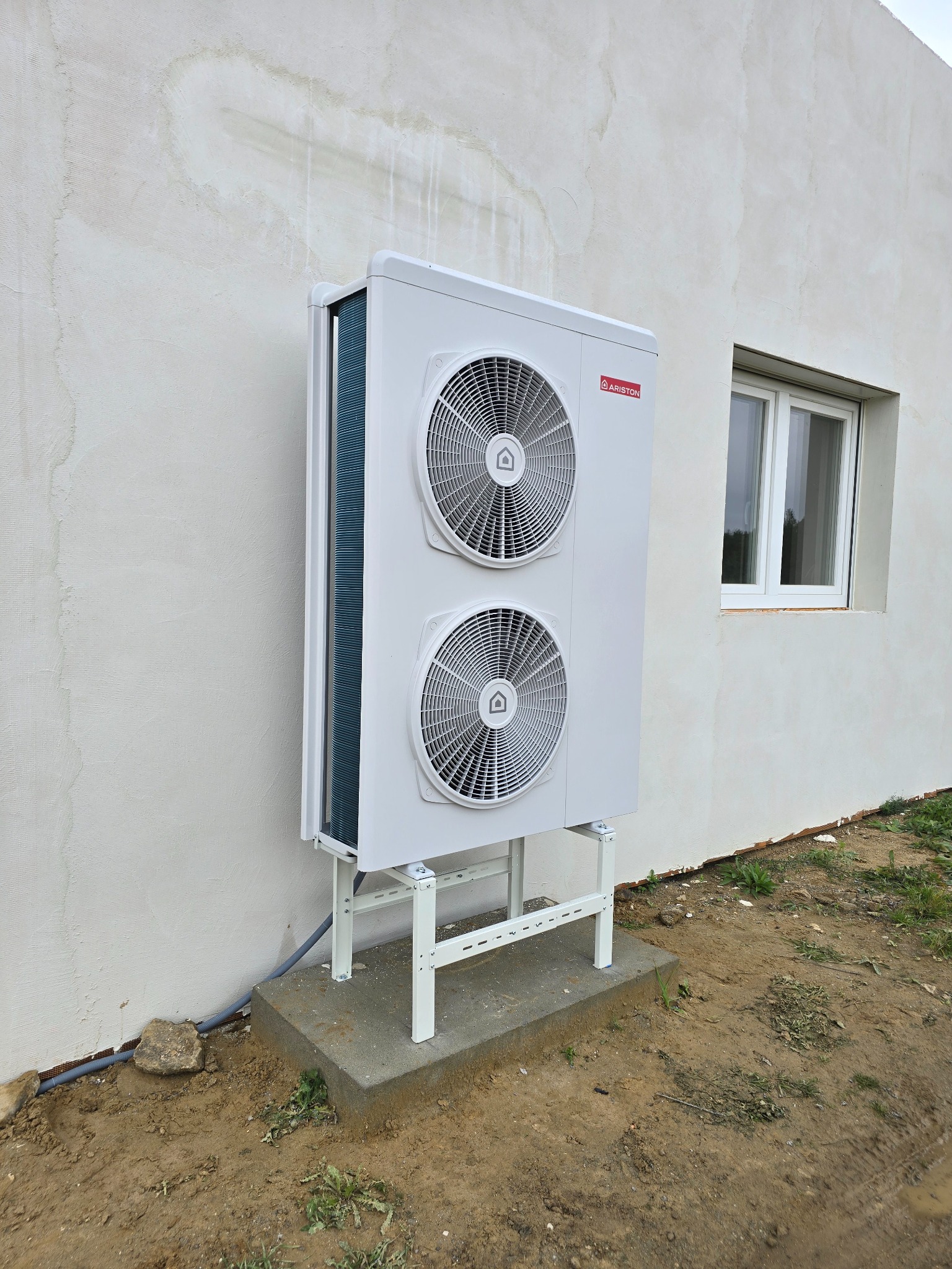 Biała pompa ciepła Ariston, zamontowana przy ścianie domu z oknem. Widoczne elementy instalacji i podstawa urządzenia. Ściana z surowym wykończeniem.