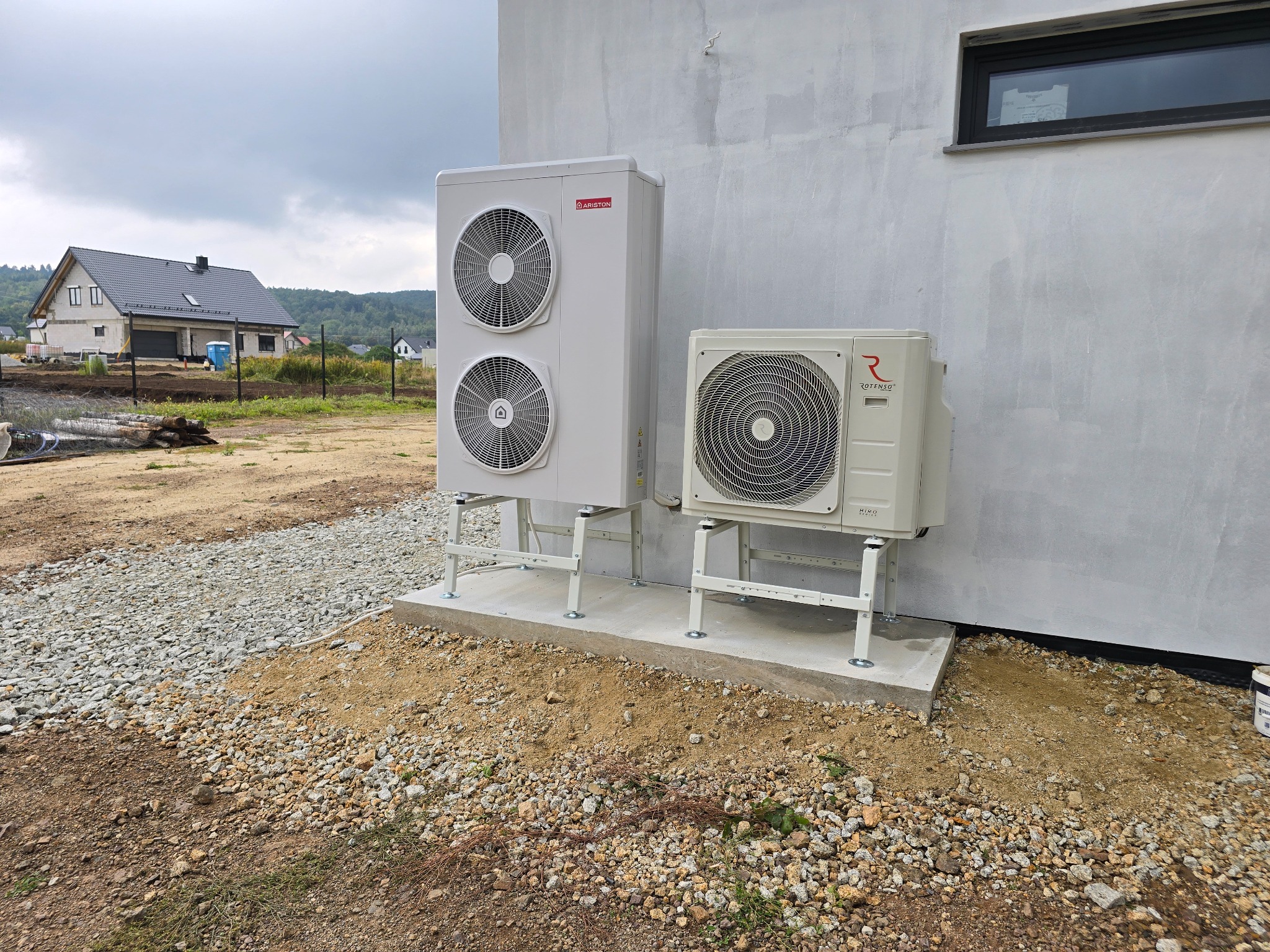 Dwie jednostki pompy ciepła (Ariston i Rotenso) zamontowane na betonowym fundamencie przy ścianie budynku. Widoczne otoczenie placu budowy i dom w tle.