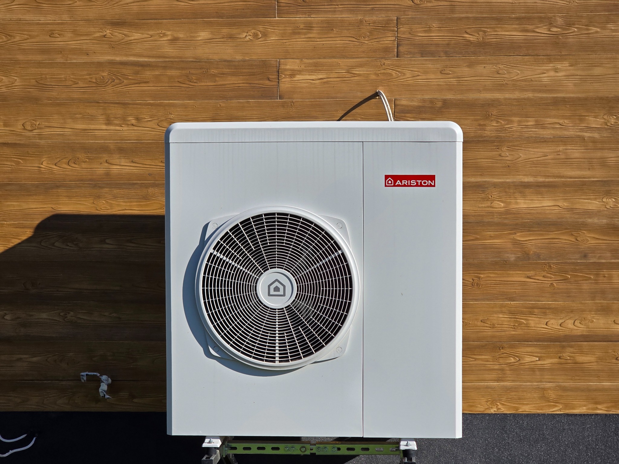 Biała pompa ciepła Ariston zamontowana na zewnątrz budynku z drewnianą elewacją. Widoczny wentylator i logo producenta. Ujęcie z bliska.