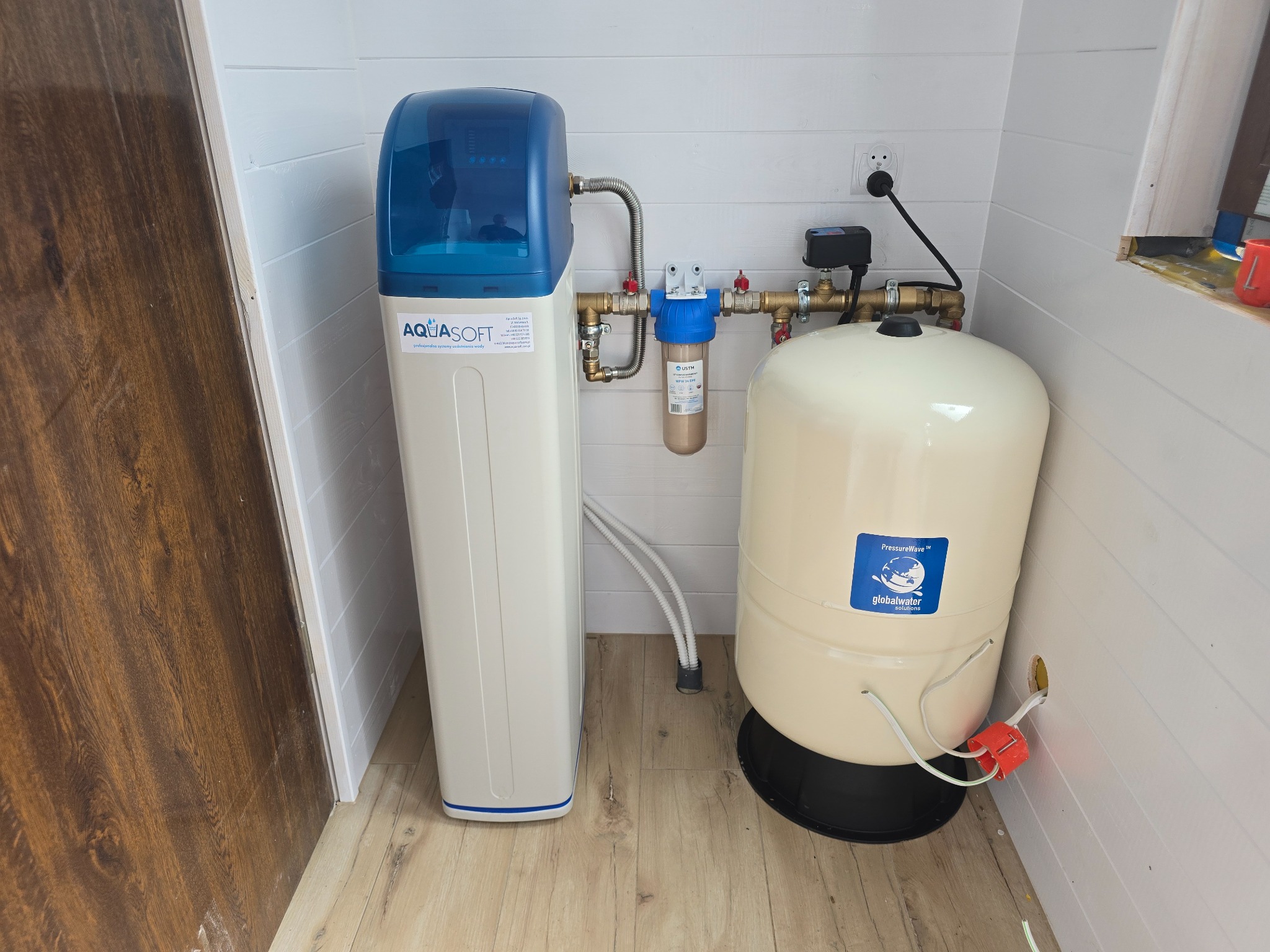Zestaw uzdatniania wody Aquasoft z zbiornikiem ciśnieniowym Global Water w białym pomieszczeniu z boazerią. Widoczne rury, filtry i przewody elektryczne.