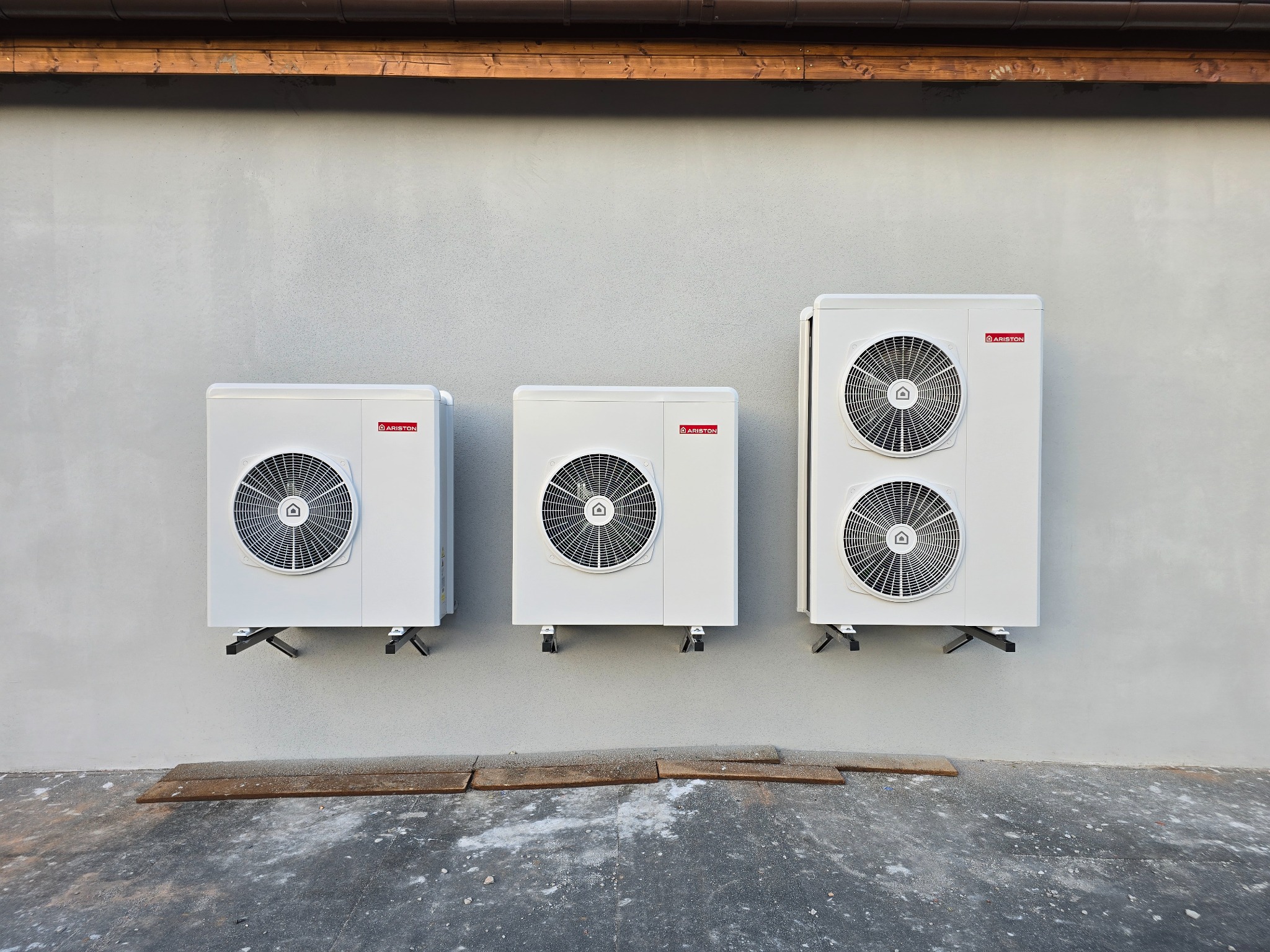 Trzy jednostki zewnętrzne pompy ciepła Ariston zamontowane na szarej ścianie budynku, widoczne wsporniki montażowe i fragment drewnianego zadaszenia.