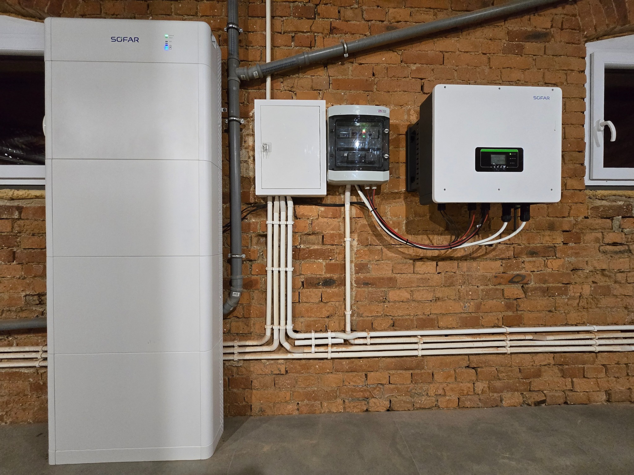 Instalacja pompy ciepła SOFAR na ceglanej ścianie. Widoczne białe moduły, skrzynka elektryczna i okablowanie. Elementy instalacji w pomieszczeniu z oknem.