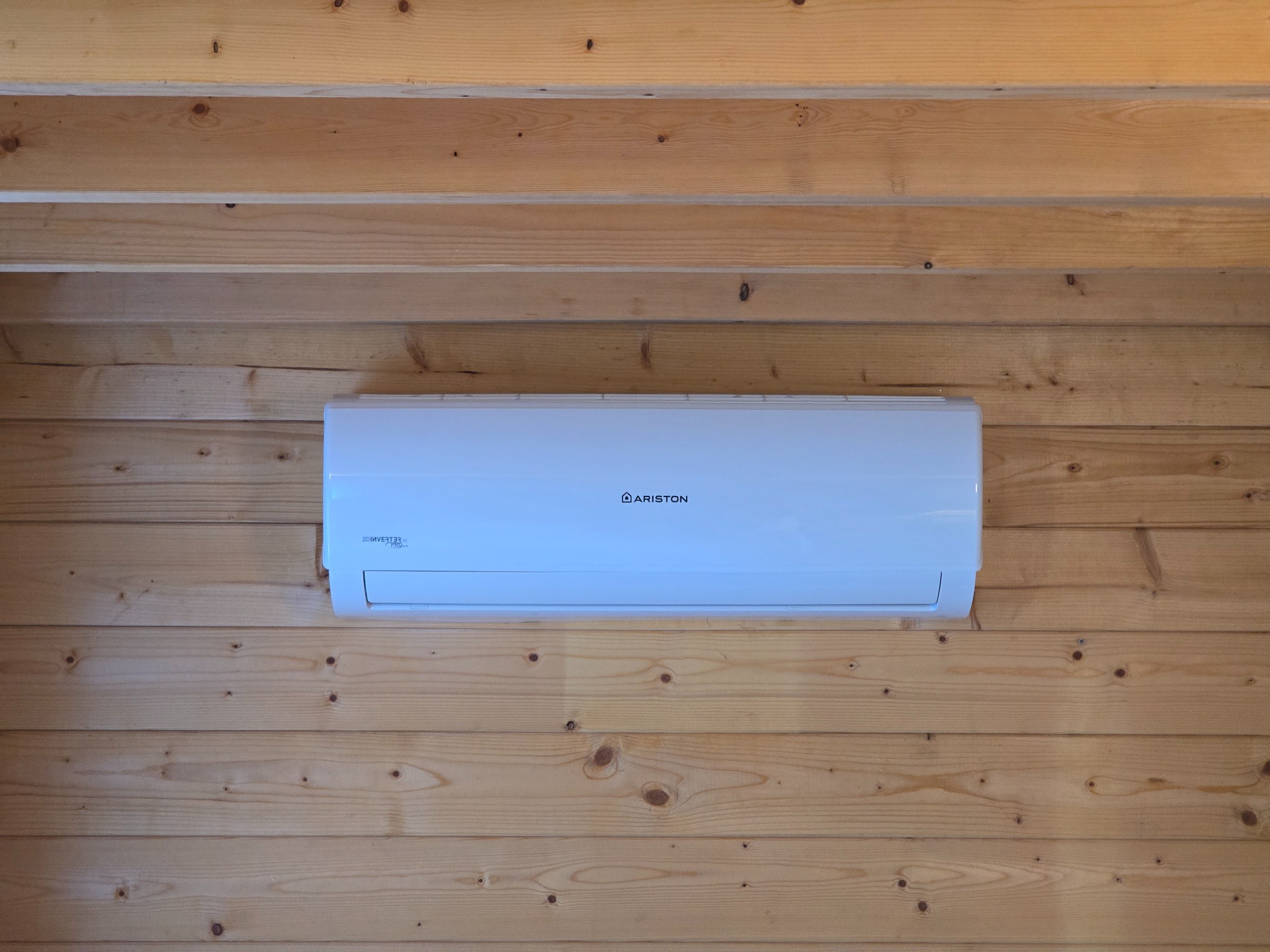 Biała klimatyzacja Ariston zamontowana na ścianie z jasnego drewna wewnątrz pomieszczenia. Widoczne słoje drewna i detale urządzenia.