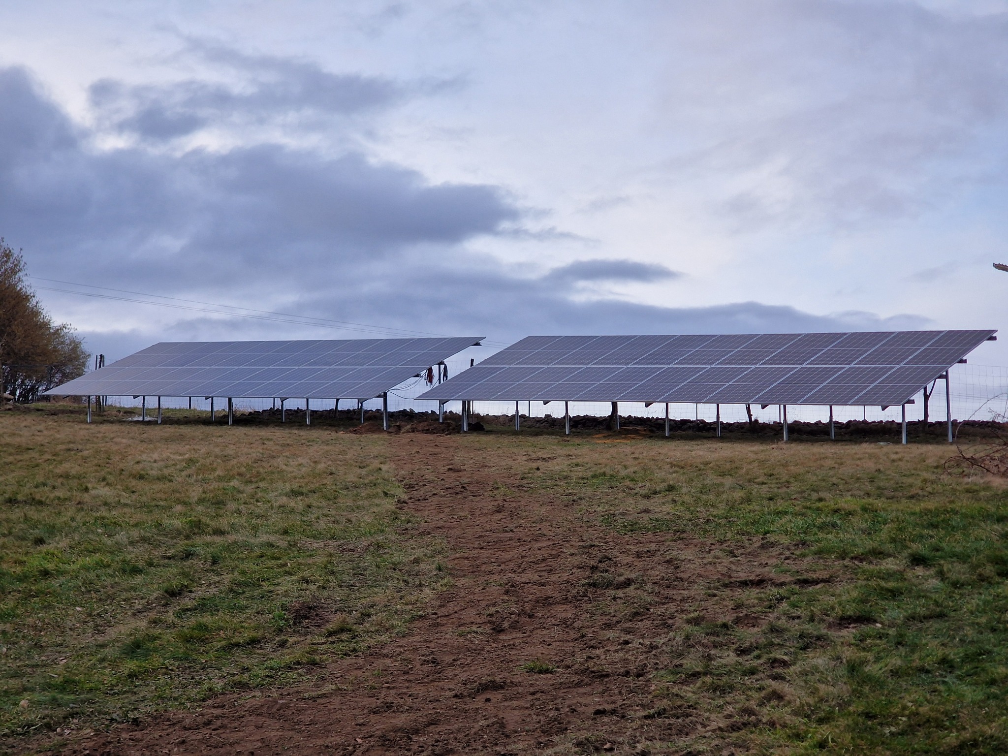 Dwa rzędy paneli słonecznych na metalowych konstrukcjach, ustawione na nierównym terenie trawiastym pod pochmurnym niebem. Ślady ziemi prowadzą do instalacji.