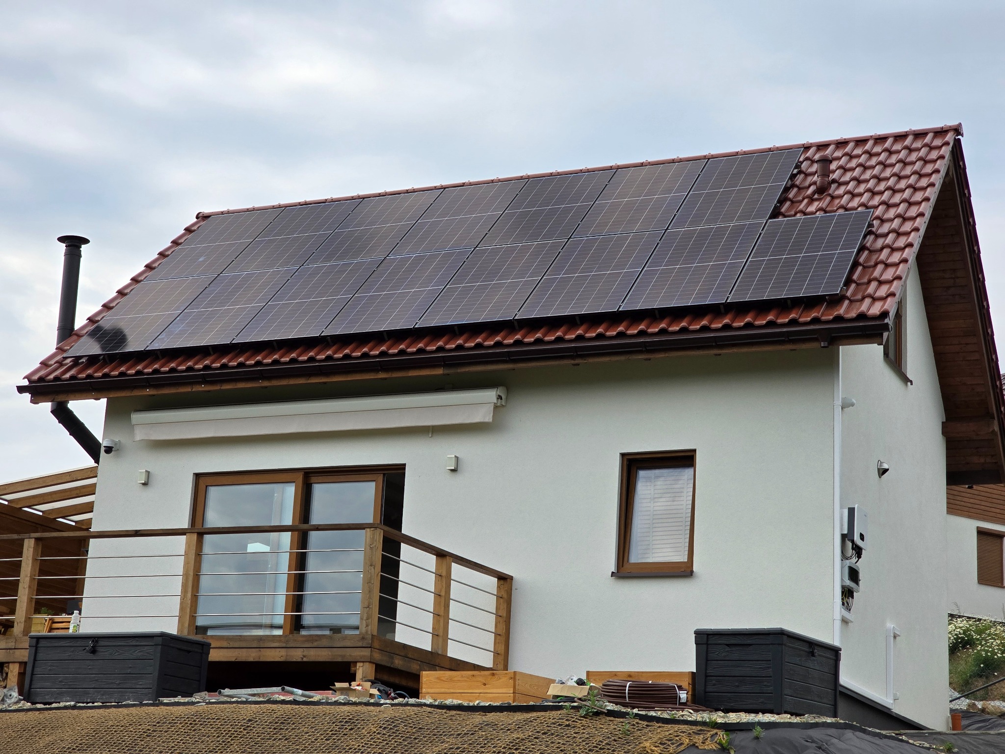 Biały dom z brązowym dachem i panelami słonecznymi. Widoczny balkon z drewnianą balustradą i czarne skrzynie. Pochmurne niebo w tle.