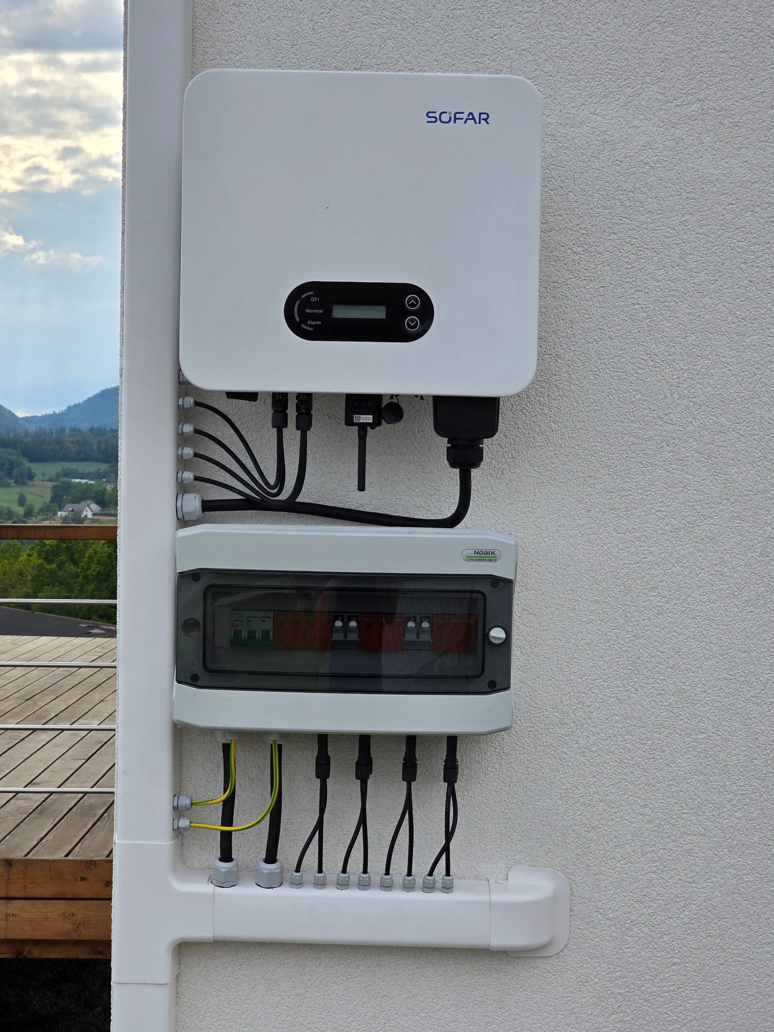 Biały inwerter SOFAR i skrzynka rozdzielcza NOARK zamontowane na elewacji budynku z widocznymi przewodami. W tle zieleń drzew i górski krajobraz.