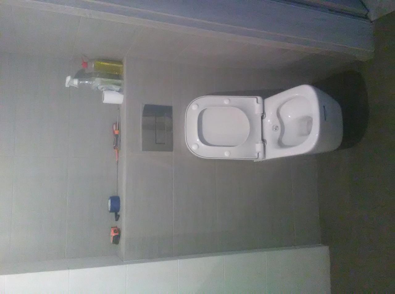 Wisząca toaleta z otwartą klapą w łazience wyłożonej szarymi płytkami. W tle widoczne przybory toaletowe i narzędzia wiszące na ścianie.