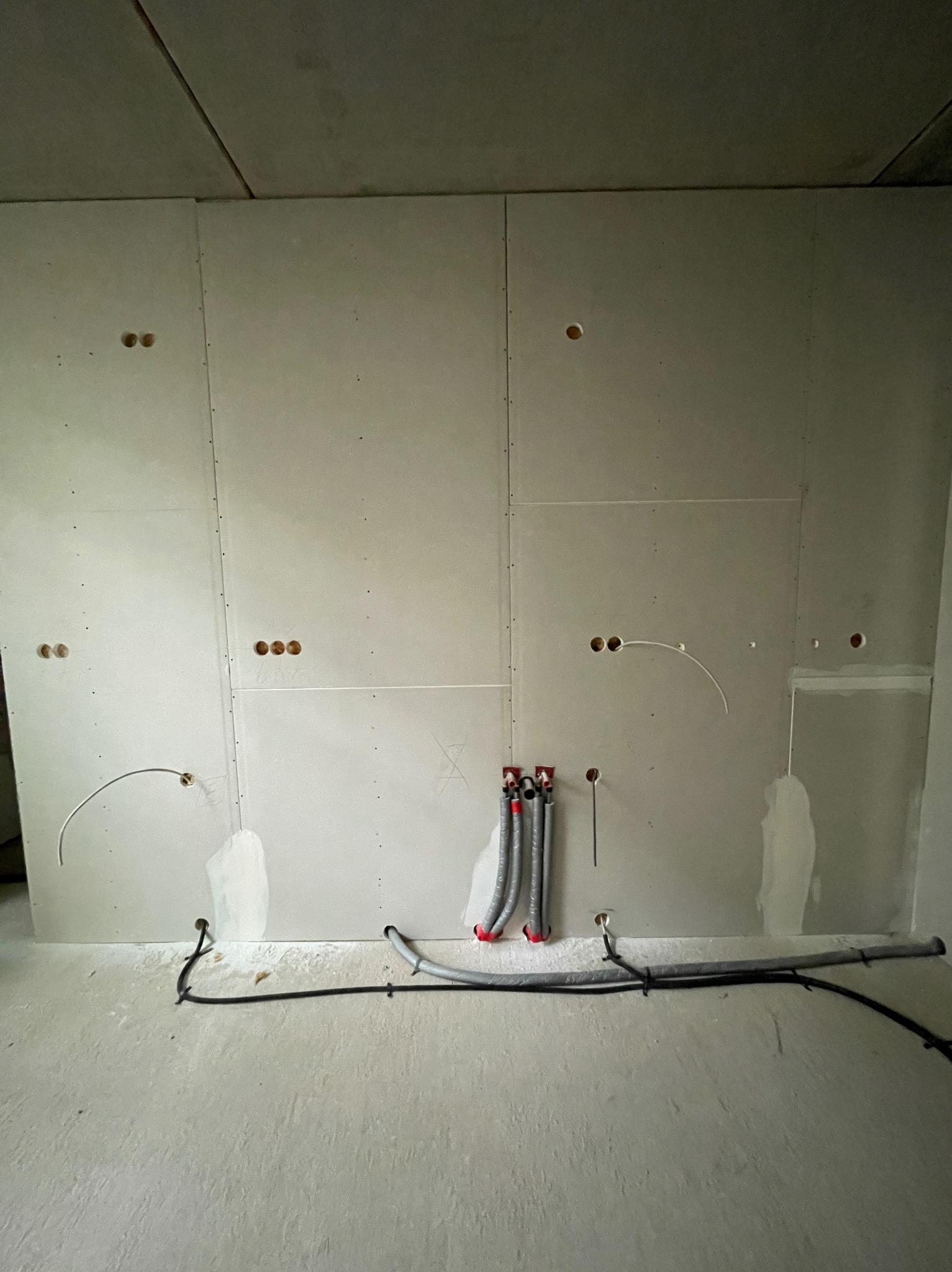 Ściana z płyt gipsowo-kartonowych w trakcie prac instalacyjnych. Widoczne otwory na gniazdka, rury w peszlach i przewody elektryczne. Surowy stan wykończenia.