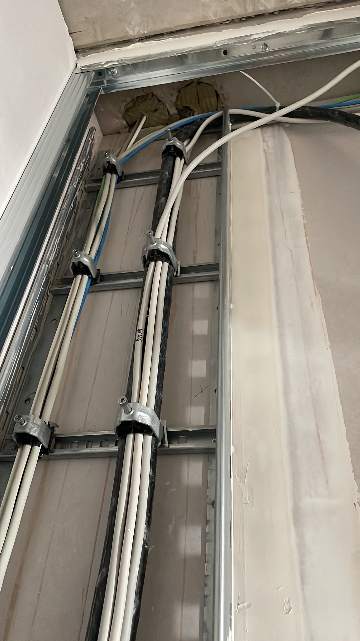 Metalowa konstrukcja z przymocowanymi kablami elektrycznymi wewnątrz ściany, widoczne otwory w ścianie z izolacją. Etap prac instalacyjnych.