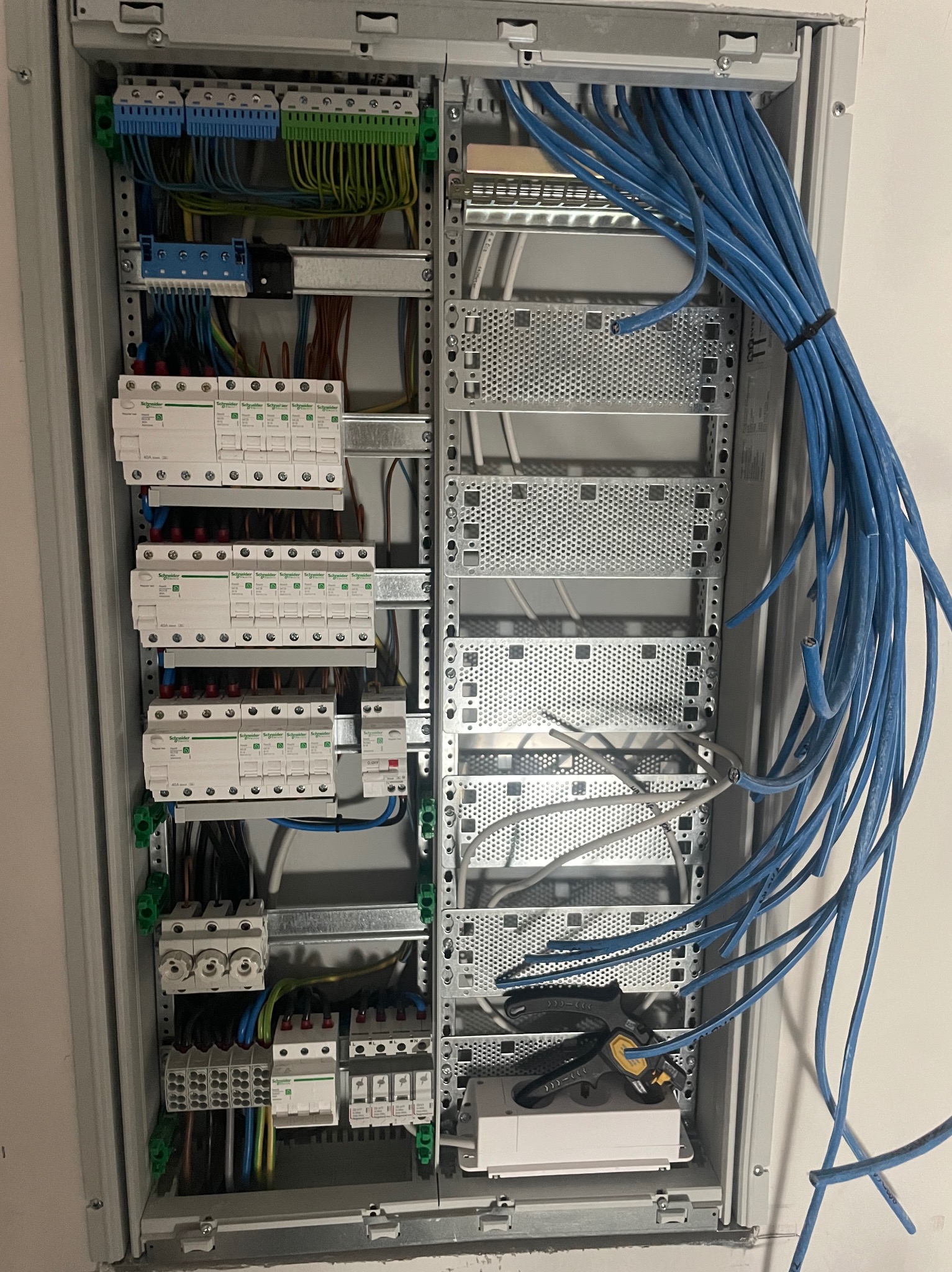 Rozdzielnica elektryczna w trakcie montażu: widoczne zabezpieczenia, listwy zaciskowe, przewody w kolorze niebieskim i narzędzia. Oświetlenie górne, kable uporządkowane.