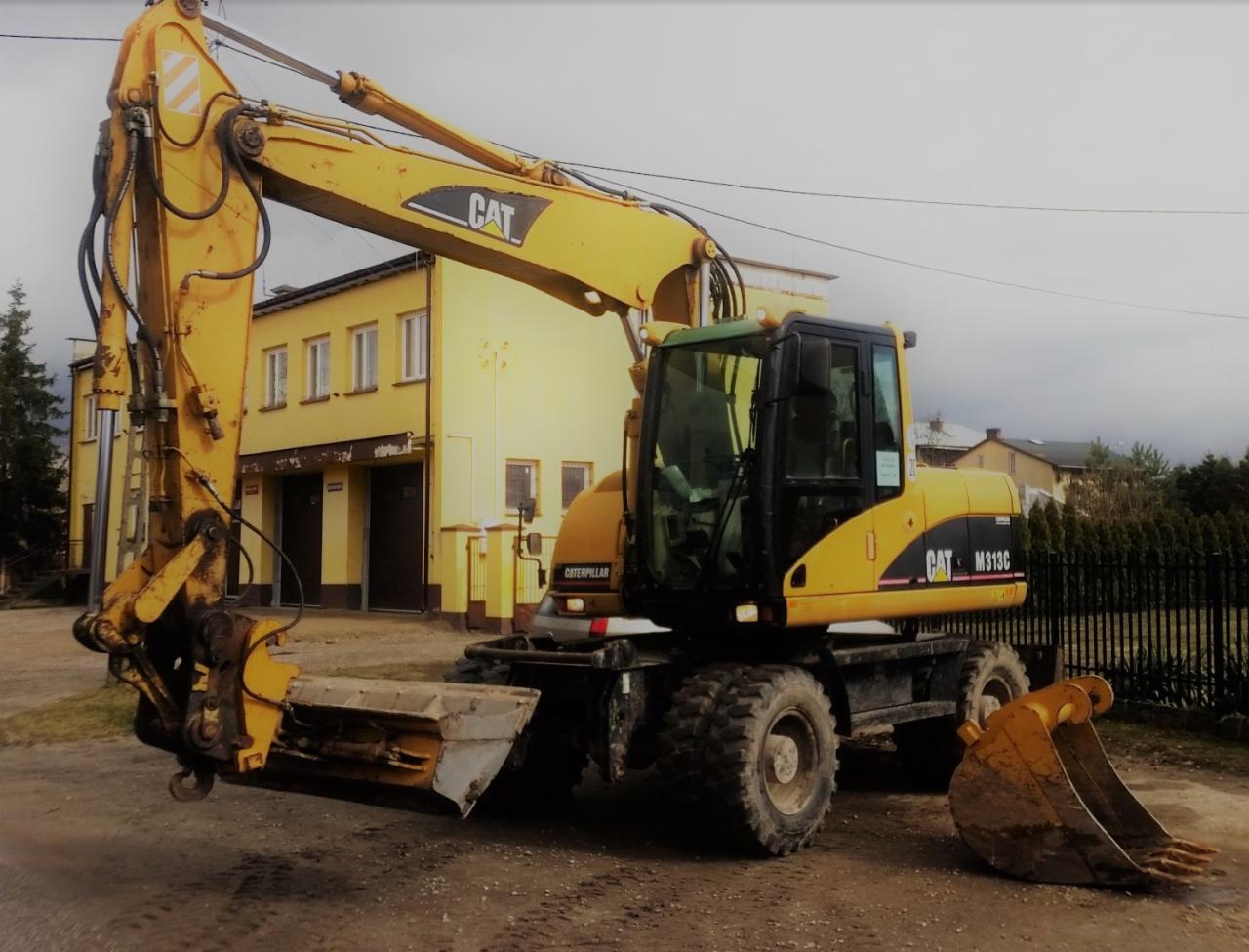 Żółta koparka kołowa CAT M313C z łyżką trapezową i łyżką kopiącą na ziemnym podłożu, na tle żółtego budynku z bramami garażowymi.