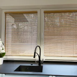 Jasne, drewniane lamele żaluzji w oknie kuchennym, z widokiem na zewnątrz, czarny zlew z wysoką baterią i biały storczyk w doniczce na blacie.