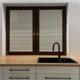 Jasne, plisowane osłony okienne w ciemnobrązowych ramach, zamontowane w oknie nad blatem kuchennym z czarnym zlewem i białymi szafkami.