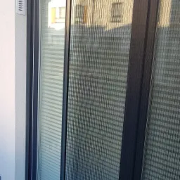 Zamknięta plisowana siatka przeciw owadom w ciemnej ramie, zamontowana na balkonie, widok na budynek za oknem.