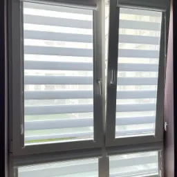 Okno z zamontowanymi roletami dzień noc w białej kasecie; widoczne naprzemienne pasy zaciemniające i transparentne.