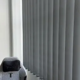 Szare żaluzje pionowe zamontowane w rogu pokoju, widoczne obok fotela gamingowego z zagłówkiem.