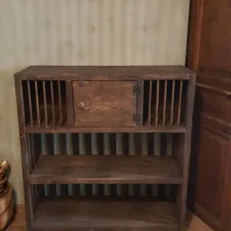 Ręcznie robiony, drewniany regał z zamkniętą szafką pośrodku, z pionowymi szczebelkami po bokach i dwoma otwartymi p&oacute;łkami poniżej, stojący na drewnianej podłodze.