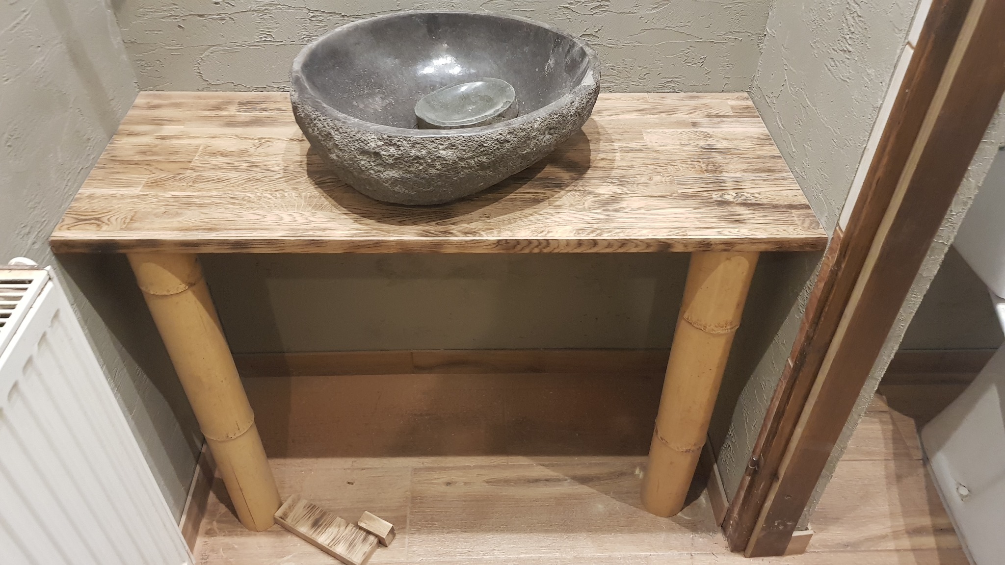 Blat drewniany z umywalką z kamienia, nogi bambusowe, widoczne fragmenty podłogi i ściany o szarej strukturze.