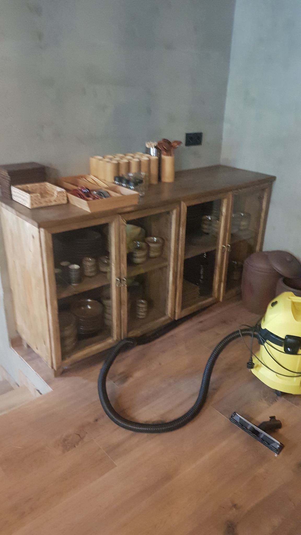 Drewniana witryna z przeszklonymi drzwiczkami, wypełniona naczyniami, obok stoi odkurzacz podczas sprzątania.