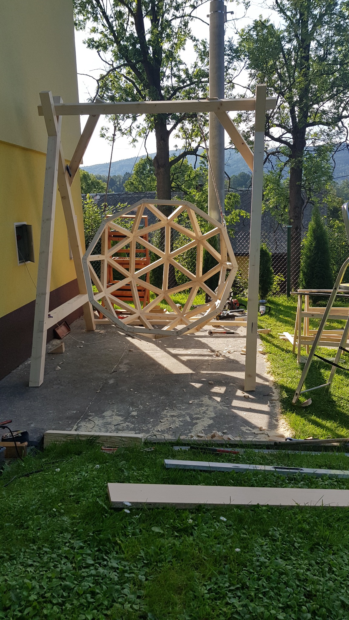 Konstrukcja drewniana w kształcie kuli zawieszona na drewnianym stelażu w ogrodzie, w tle drzewa i góry, widoczne narzędzia i materiały budowlane.