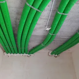 Zielone, karbowane rury wentylacyjne przymocowane metalowymi obejmami do surowego betonowego stropu, uszczelnione pianką montażową przy przejściu przez ścianę.