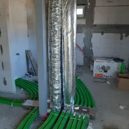 Dwa pionowe przewody wentylacyjne owinięte folią aluminiową, połączone z zielonymi, karbowanymi rurami rozprowadzonymi na podłodze w surowym wnętrzu budynku w trakcie budowy.
