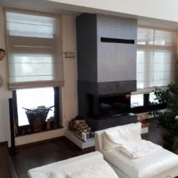 Beżowe rolety rzymskie w nowoczesnym salonie z kominkiem obłożonym szarym kamieniem, widok z wnętrza na okno z dekoracjami.