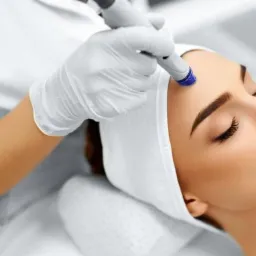 Kobieta z zamkniętymi oczami podczas zabiegu oczyszczania skóry twarzy za pomocą urządzenia z niebieską końcówką, biały turban na głowie, rękawice ochronne na dłoniach kosmetologa.